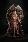 A blonde woman sits on a large metallic throne-like chair against a black studio background, wearing a red draped outfit and nude top. No visible brand markings are present.
